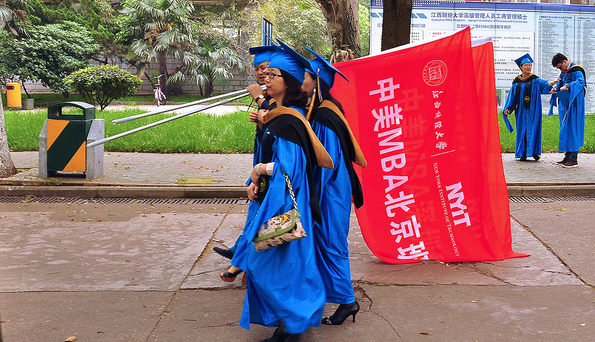  NYIT-JUFE students in cap and gowns holding a flag.