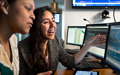 Two students talk and reference computer monitors.